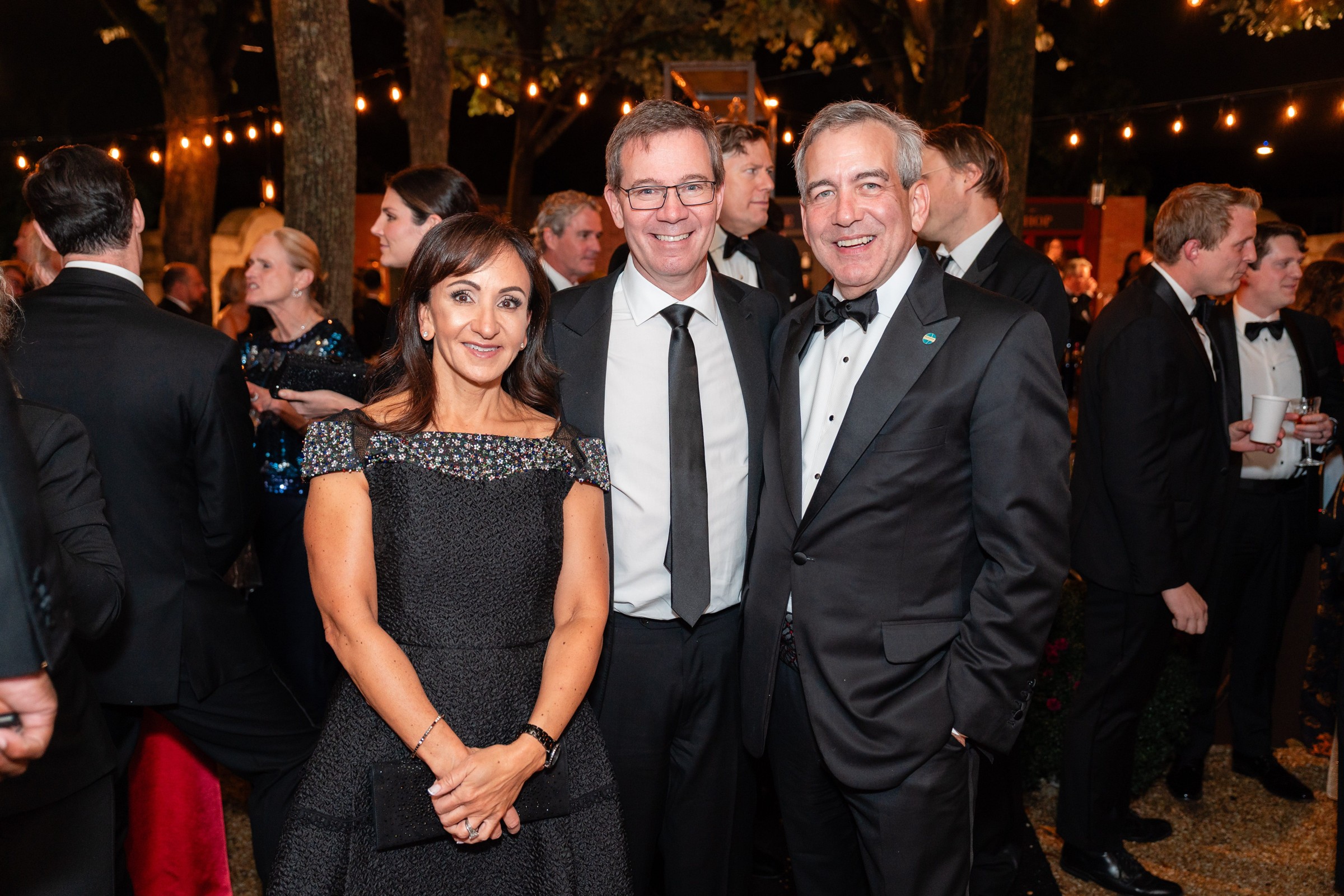 Three people in formal attire smiling together at an evening outdoor event with string lights in the background.
