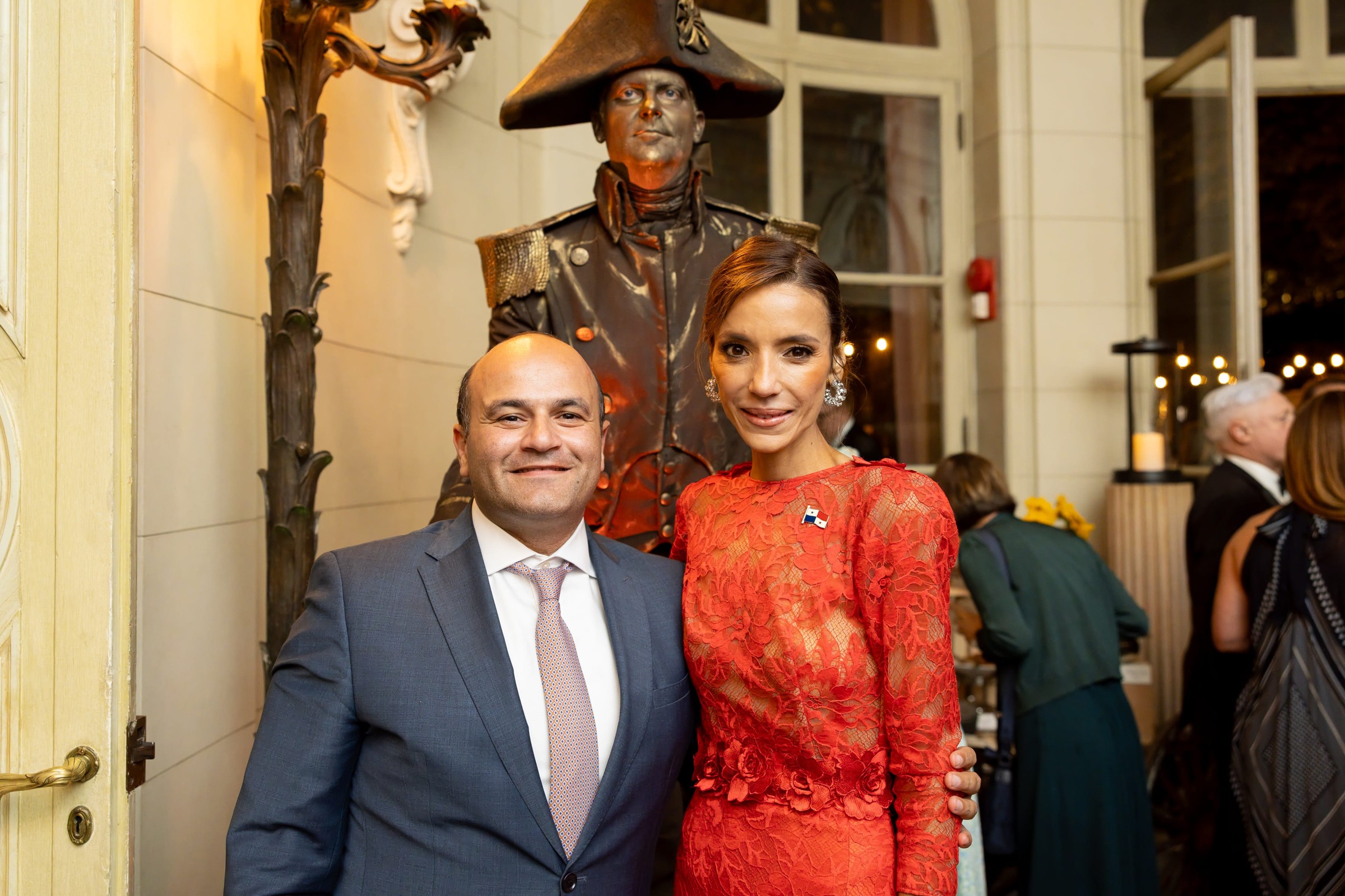 Two people posing together with a statue of a historical figure in the background at an elegant indoor event.