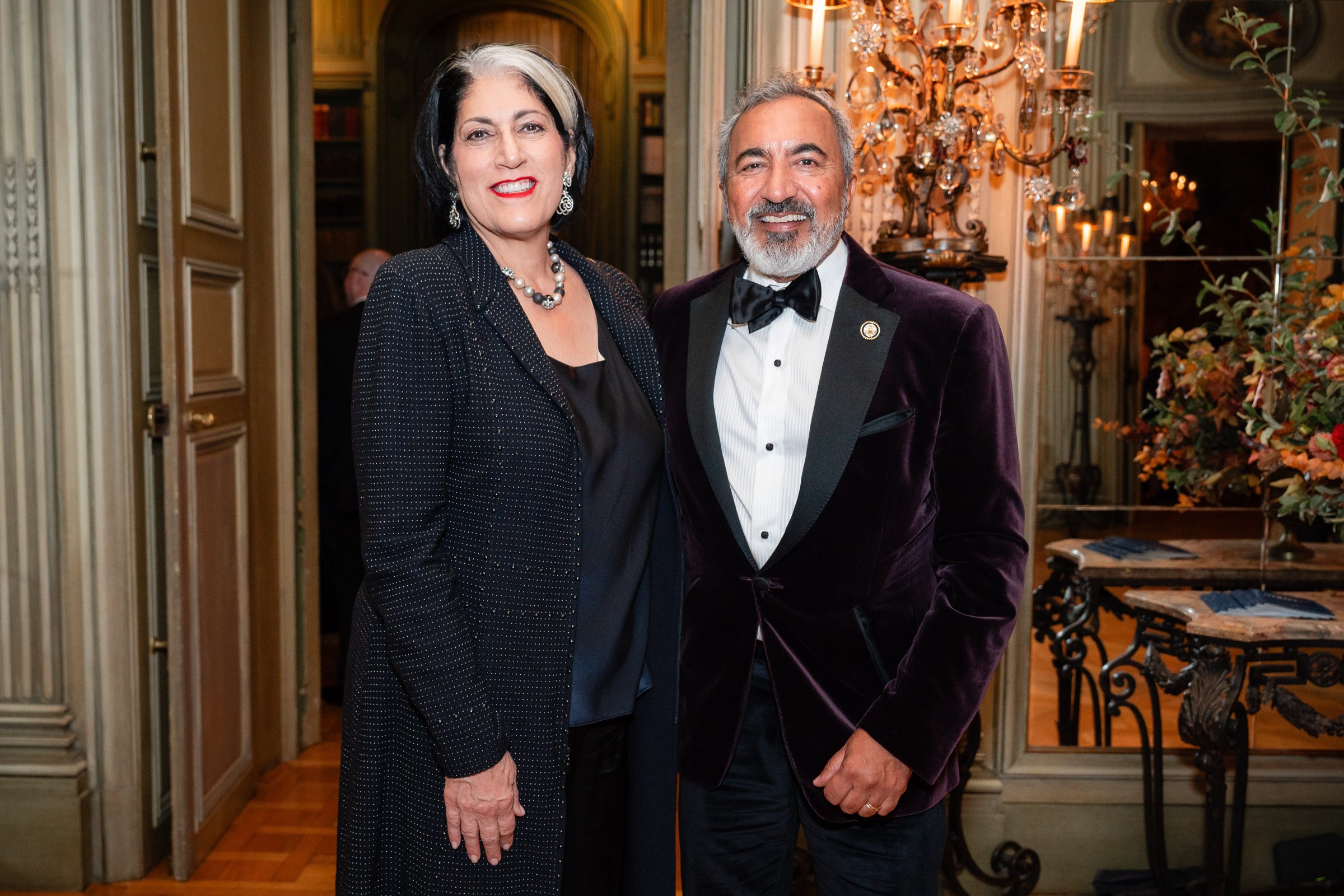 A man in a tuxedo stands beside a woman in a black outfit, both smiling in an elegant indoor setting.