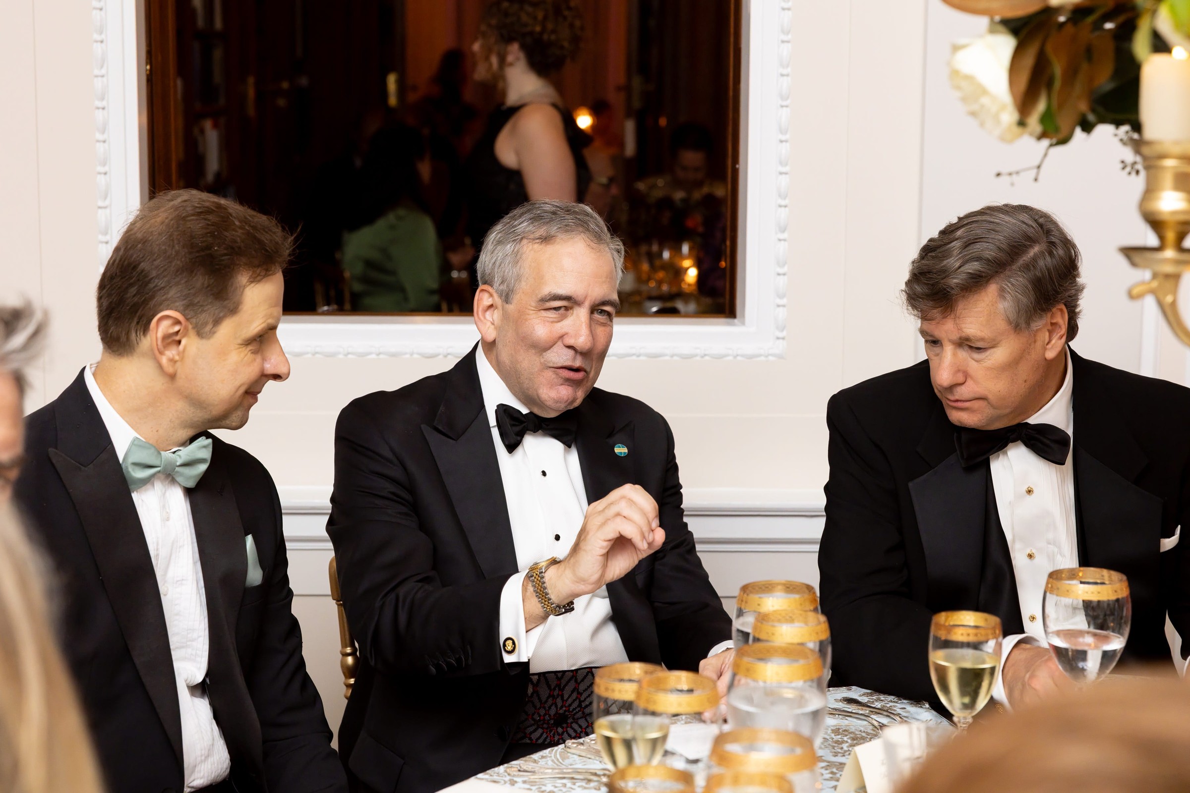Three men in formal attire engaged in conversation at a dinner table with wine glasses and decorative elements.