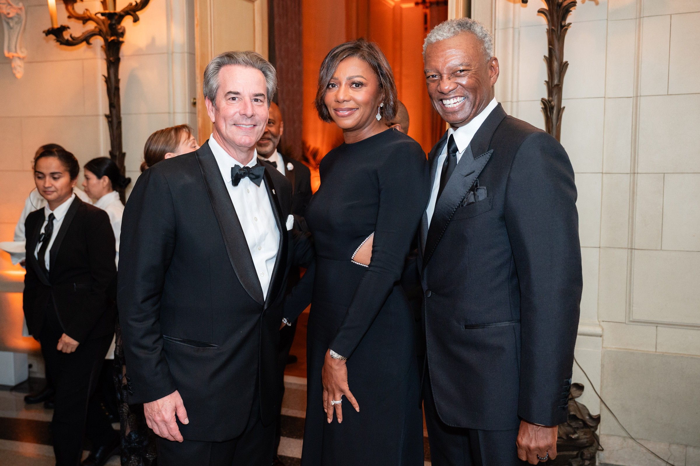 Three elegantly dressed individuals posing together at a formal event, with staff serving in the background.