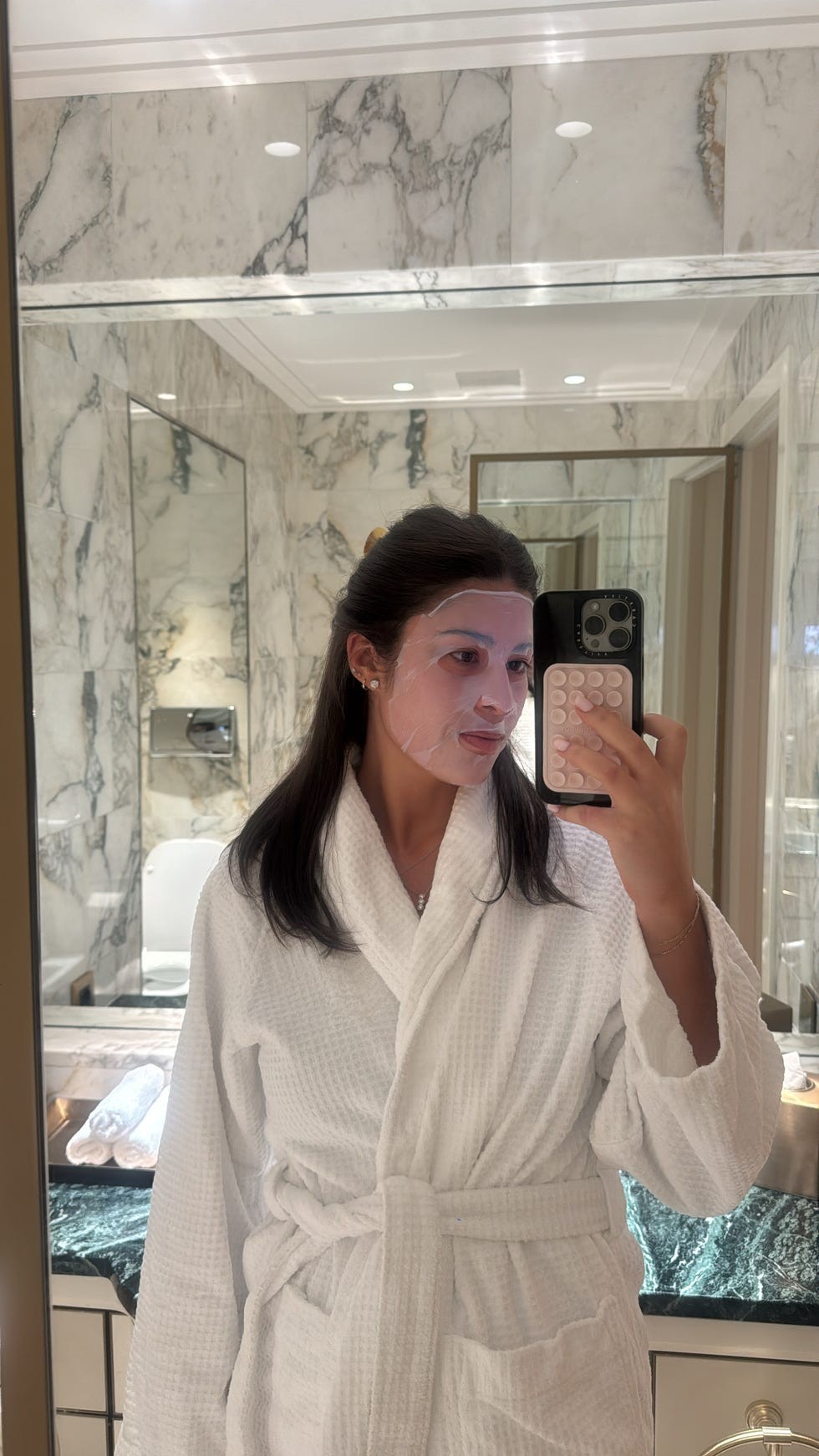 A person wearing a facial mask and white robe, taking a selfie in a luxurious bathroom with marble walls.