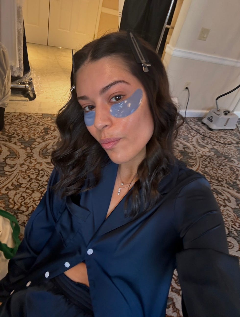 Person with a blue under-eye mask, wearing a dark shirt, posing in a room with patterned carpet and a doorway in the background.