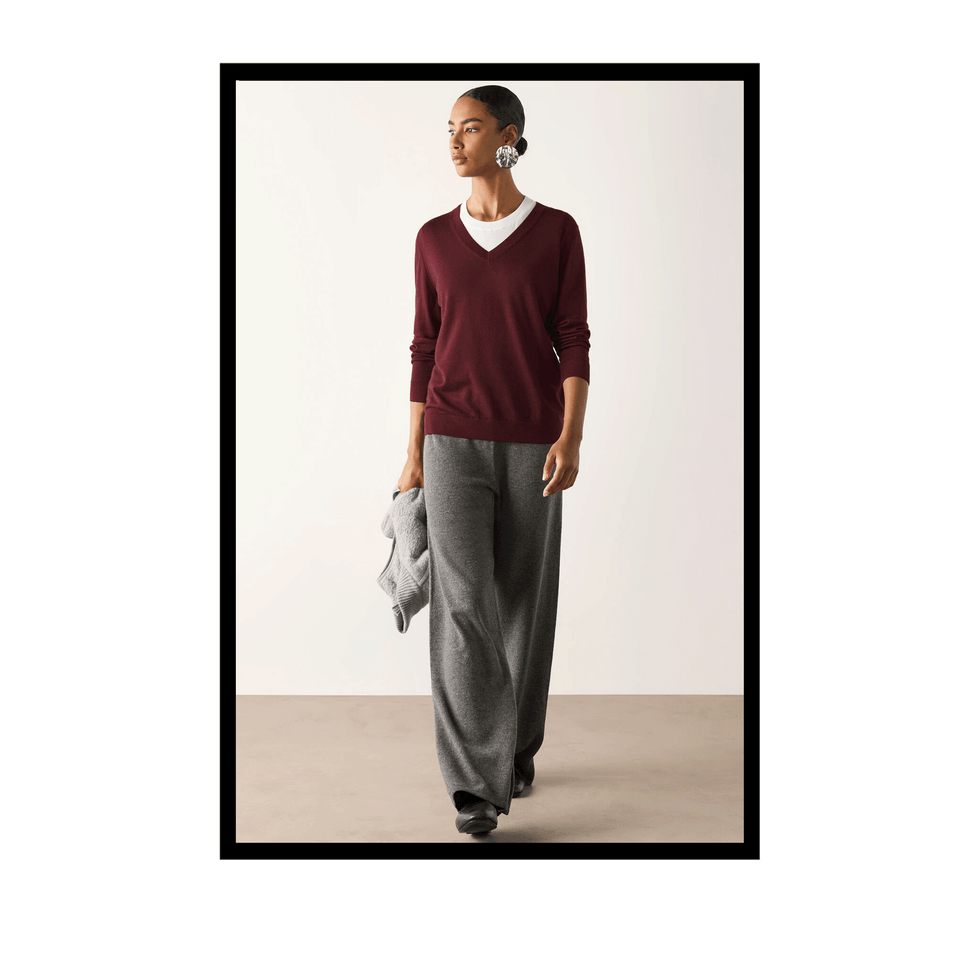 Model wearing a burgundy sweater and gray wide-leg pants, holding a gray sweater on a neutral background.