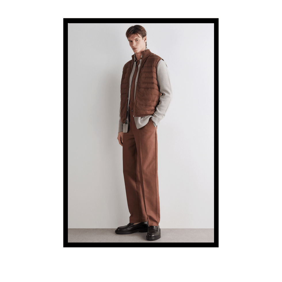 Young man in a brown quilted vest, grey shirt, and brown trousers, standing against a plain wall.