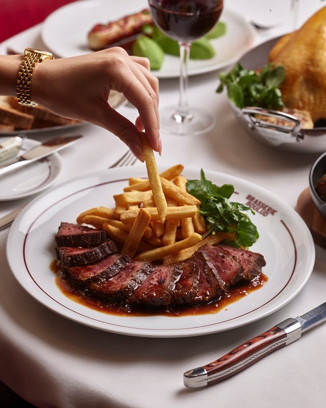 A hand reaching for a French fry next to sliced meat and greens on a dining table with a glass of wine in the background.