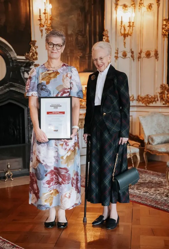 Two women standing in a lavish room, one holding a certificate, dressed in elegant attire.