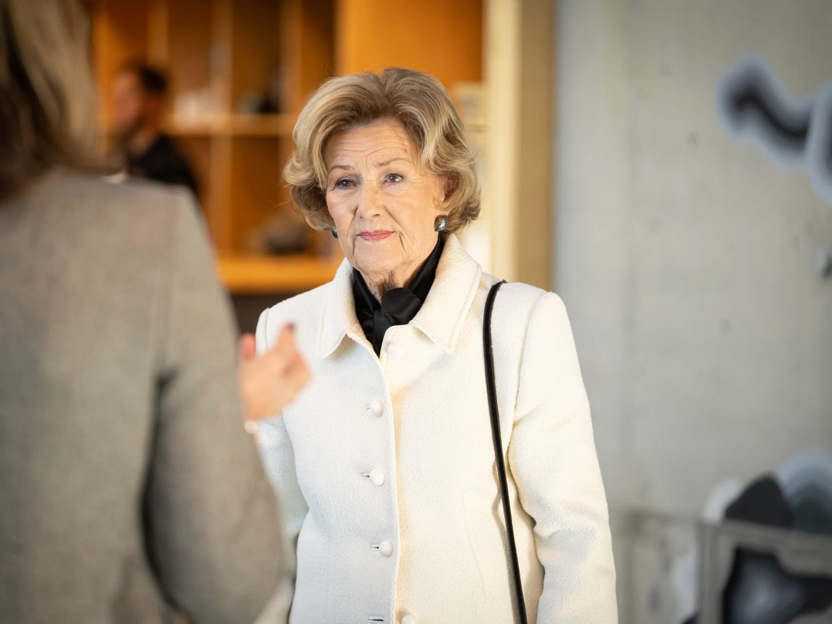 A woman in a white coat listens intently during a conversation in a modern setting.