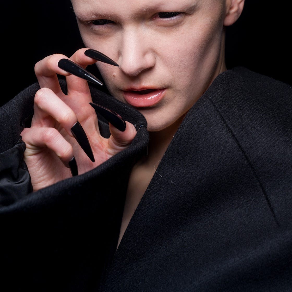 A person with a bald head and long, sharp black nails poses dramatically against a dark background.