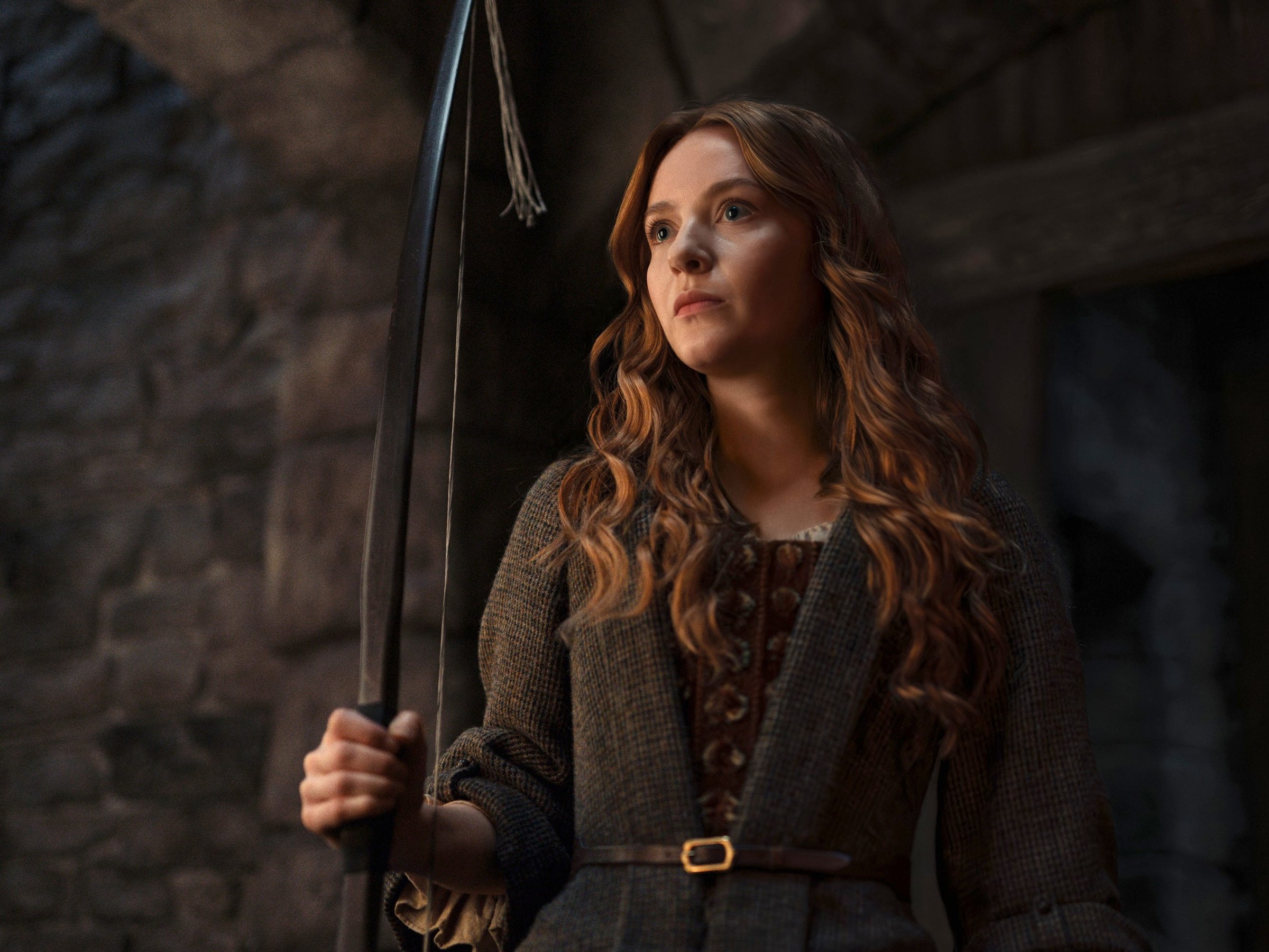 Young woman with long hair holding a large scythe, dressed in a medieval-style outfit, standing in a dimly lit stone setting.