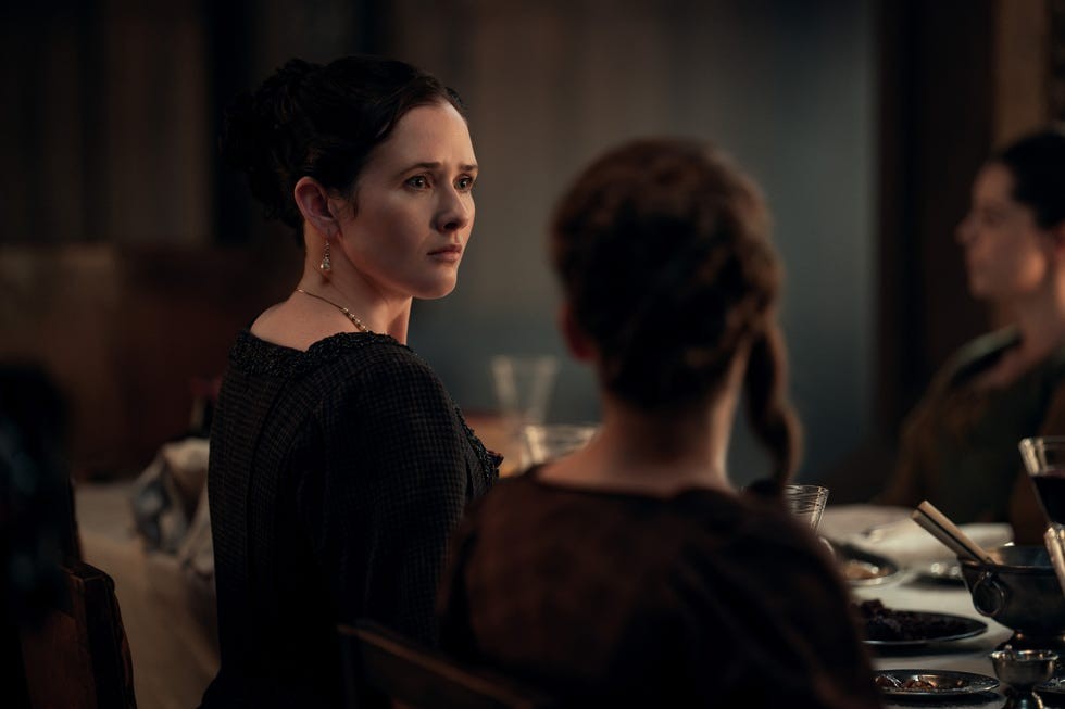 A woman with an anxious expression seated at a dimly lit dinner table, looking towards others.