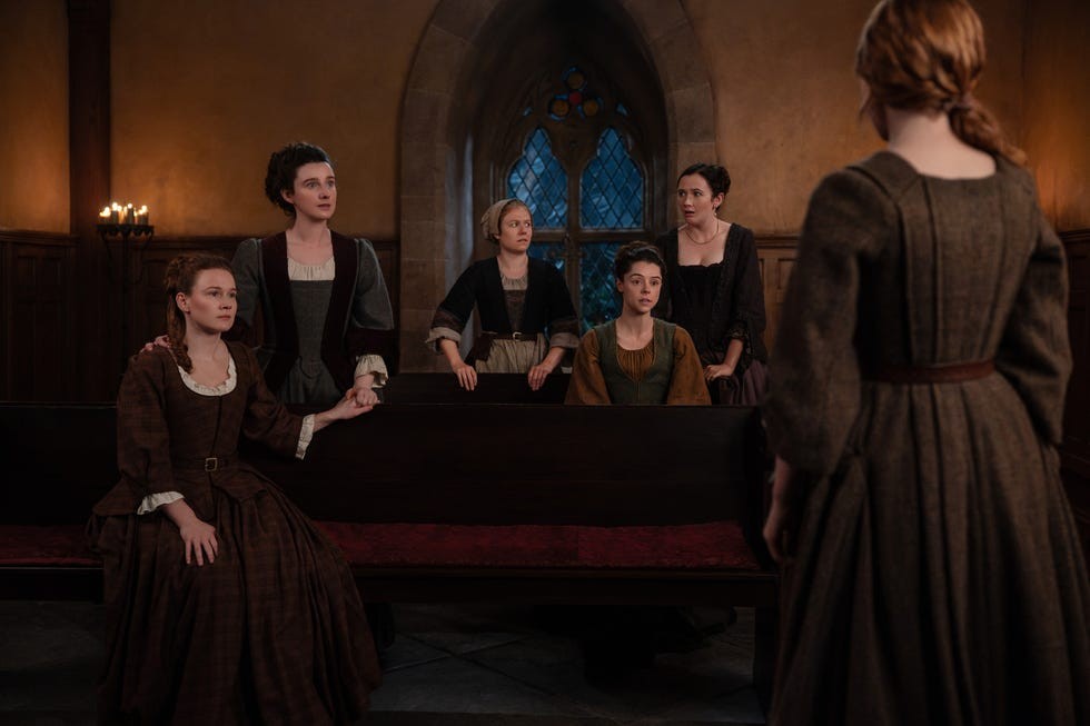 A group of women in historical attire seated in a dimly lit room, engaged in an intense discussion.