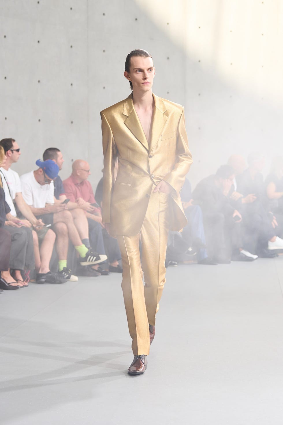 Model in a shiny golden suit walking on a runway with a crowd seated in the background.