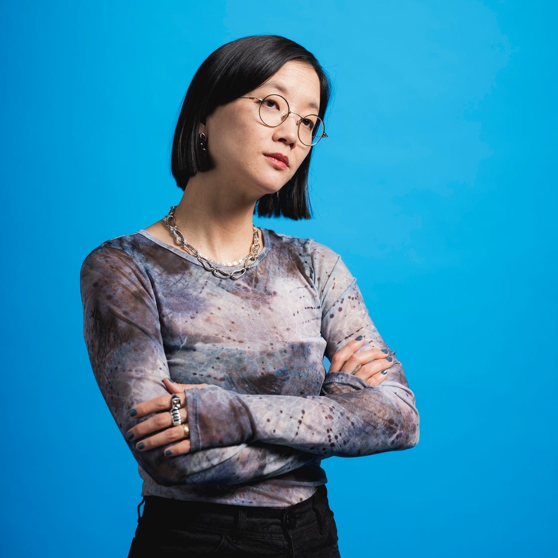 Person with short black hair and glasses, wearing a patterned long-sleeve shirt, standing against a blue background.