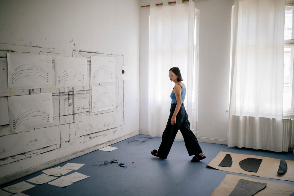 A person walking in a bright art studio with sketch papers displayed on the wall and flooring.