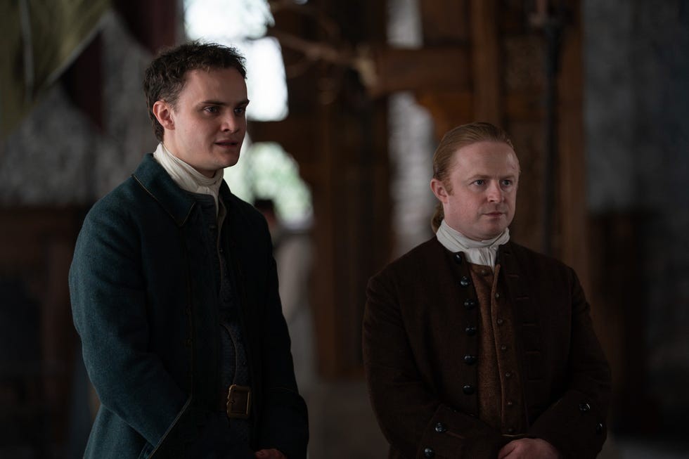Two men in historical attire standing indoors, engaged and attentive in a conversation or event.
