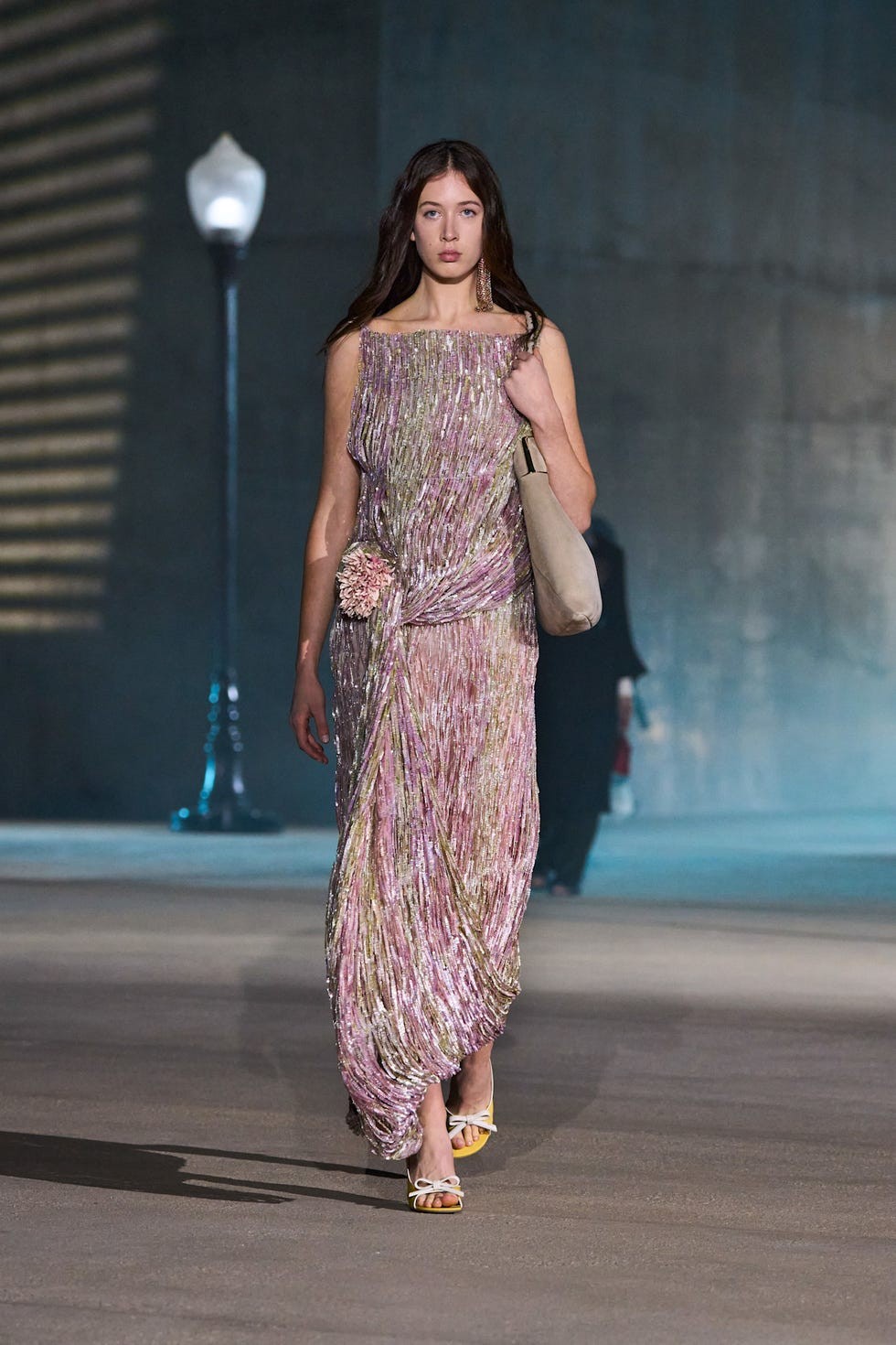 Model walking on a runway wearing a shimmering pink dress and holding a beige bag, with street lamps in the background.