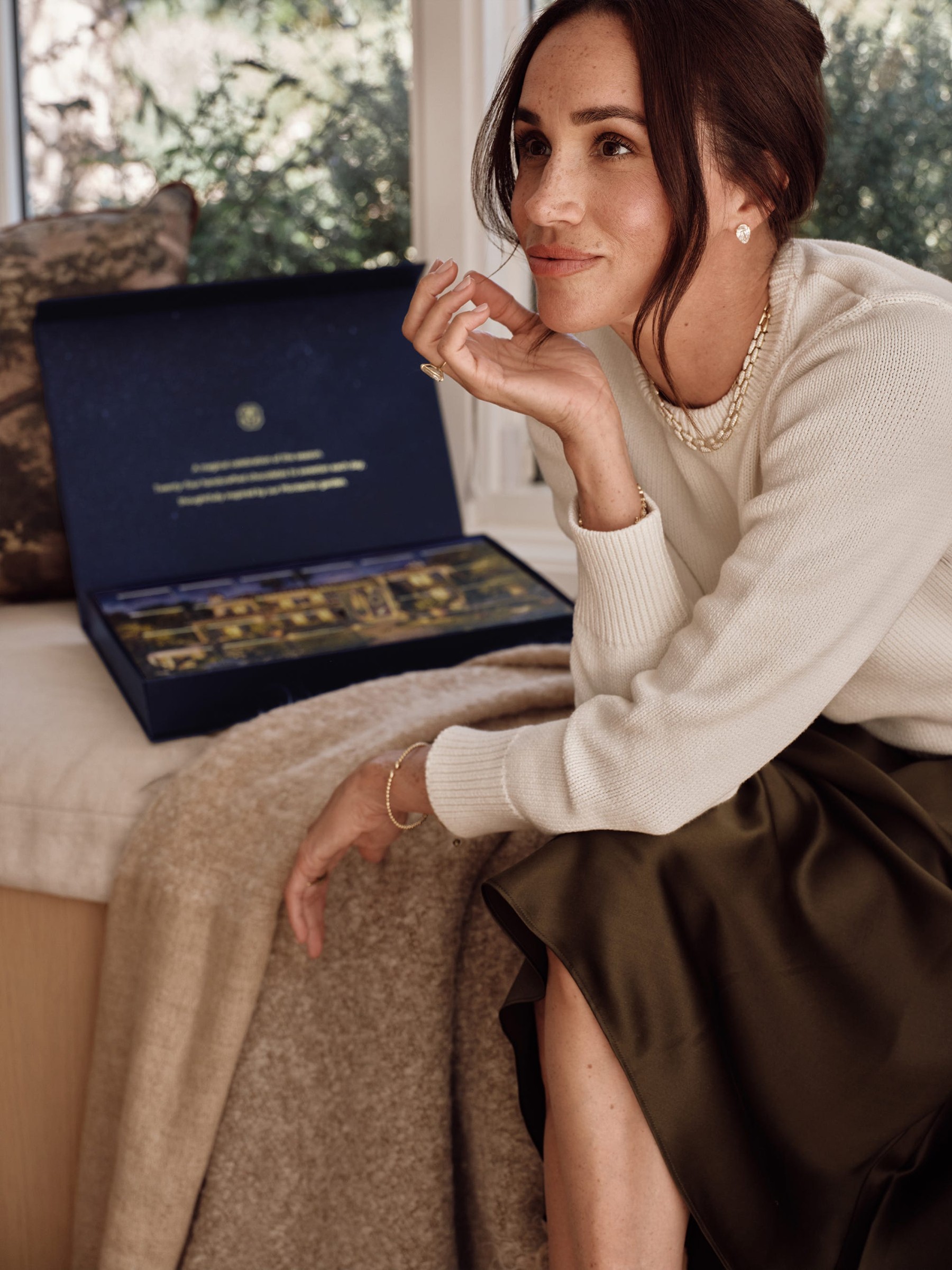 Woman in a cream sweater and green skirt sitting on a couch, thoughtfully looking at a product box nearby.